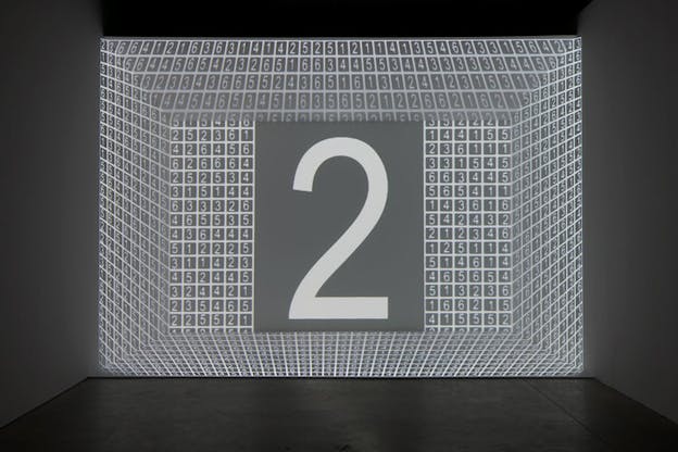 At the far wall of a dark grey room, a grid filled with numbers, 1, 2, 3, 4, and 5 is projected in such a way as to appear 3D. A large number 2 is in the middle. 