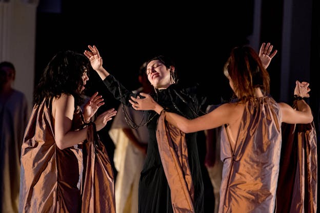 Performers in brown robes surround a figure clad in black. Their hands elevated and their eyes closed.