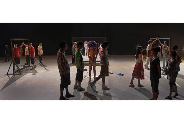 A performance still of numerous figures situated around a dimly lit space. Within the space there are numerous standing mirrors the size of humans. A few performers stand beside the mirrors, touching them. The rest of the performers stand around the space in various street clothes with their side to the audience. A few performers appear mid-stride. In the middle, one performer stands out, wearing nothing but striped shorts and a mask of a purple create with a red bandana over their head. A performance still of numerous performers in various street clothes standing in a semi-circle around a person who appears to have fallen off their bike. This performer lays on their side and by their head, there is a white framed picture. To the side of them, many bronze colored, shiny wet snake like objects are scattered on the floor.  