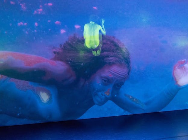 A close up performance still of nia love swimming nude, pressed up against glass. Only her face, chest, and arms are visible. The scene is dappled pink and purple. A small light green image of the back of a person upright in fetal position is positioned by love's hair. 