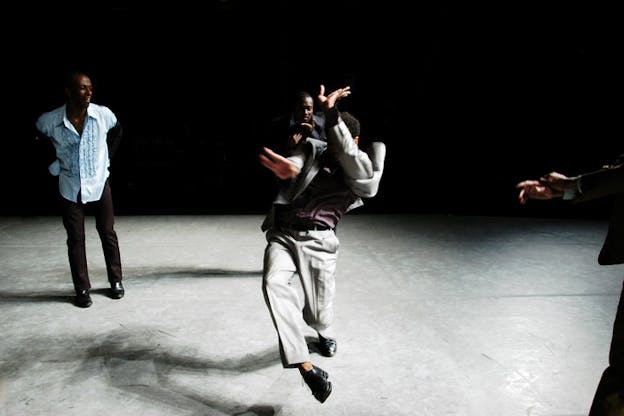 A person dressed in a gray suit dances and twists their body in the center of a black stage with gray floors while a person standing in the back left smiles and watches. 