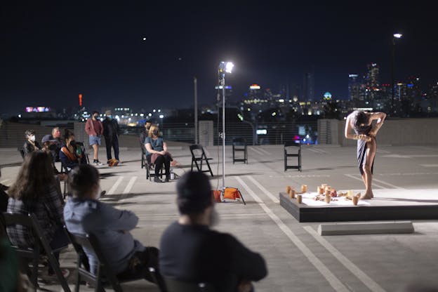 Angel Lartigue stands on a long, raised platform in a parking lot. Her torso is twisted down to her left as she lifts her purple dress to reveal her upper thigh. On the stage in front of her are clay pots. In front of the stage there are seated viewers. Behind her is a city skyline and the night sky.