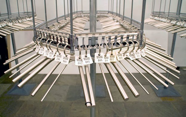 Installation view of an empty coat check rack filled numerous silver hangers with numbered tags on them. The floor is made of slate grey tiles and the walls are white. 