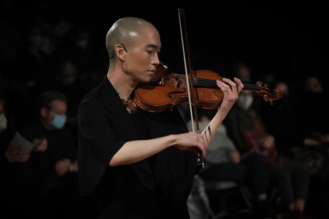 eddy kwon plays her violin, her bow poised on the strings, her eyes looking down and to the left. Her head is shaved and she is dressed in black with a microphone headset and medium hoop earrings.