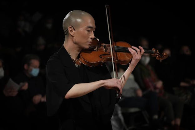 eddy kwon plays her violin, her bow poised on the strings, her eyes looking down and to the left. Her head is shaved and she is dressed in black with a microphone headset and medium hoop earrings.