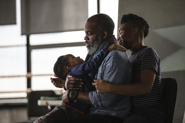 A young person sits behind and engulfs an older man in the same chair, while the second embraces a toddler. 