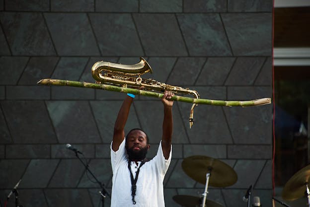 A photograph of JJJJJerome Ellis performing, closing his eyes and holding a saxophone and natural reed above his head. He stands in front of a dark blue geometric-patterned wall.