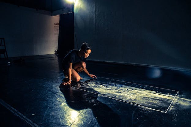 A person kneels in front of a rectangular shape on the floor with white dust scattered around it. 