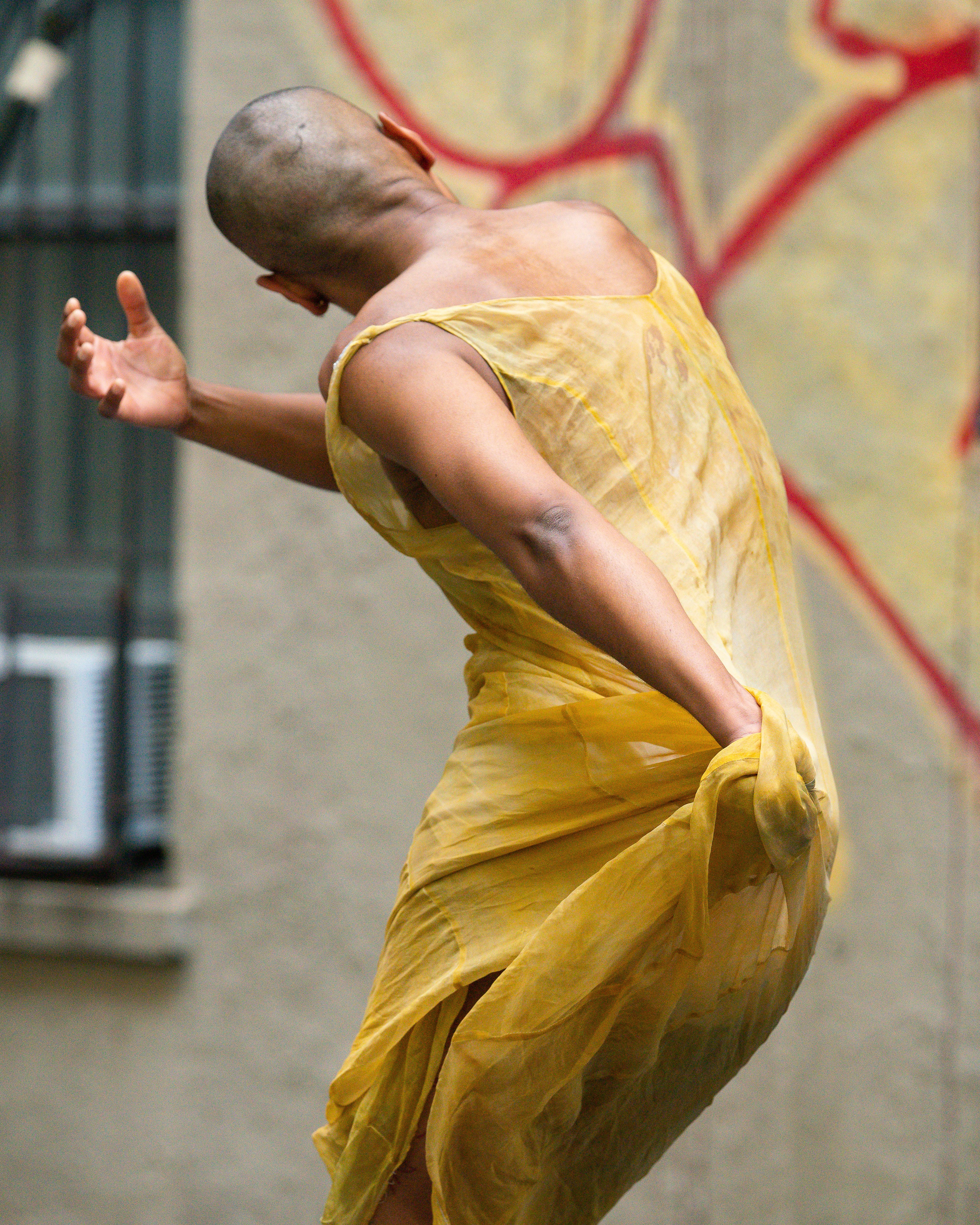 Jasmine Hearn in mid-movement wearing a bright yellow, loosely fitting dress, their back to the camera. Their torso bends and twists to the left, their hips thrust out to the right. Their right hand wraps around their body while their left hand gathers their dress behind them. Their back and shoulders glisten.