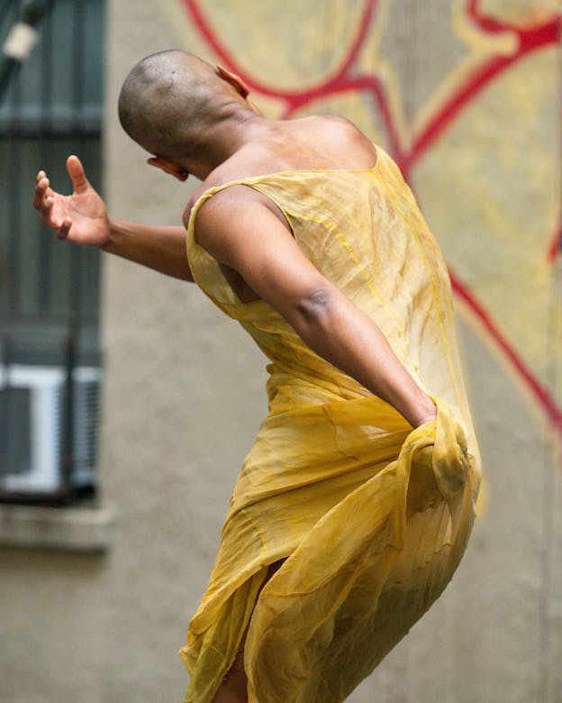 Jasmine Hearn in mid-movement wearing a bright yellow, loosely fitting dress, their back to the camera. Their torso bends and twists to the left, their hips thrust out to the right. Their right hand wraps around their body while their left hand gathers their dress behind them. Their back and shoulders glisten.