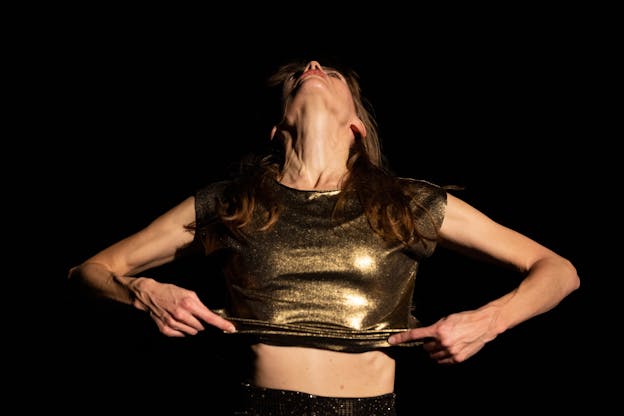Joanna Kotze performs alone on a dark stage, illuminated softly from the front. A closeup shot, she stands visible from the waist up with her body facing the camera. Her head is tilted back and she stares at the ceiling. Her hands hold and lift the hem of her gold metallic, shortsleeved top, index fingers point inward towards each other. 