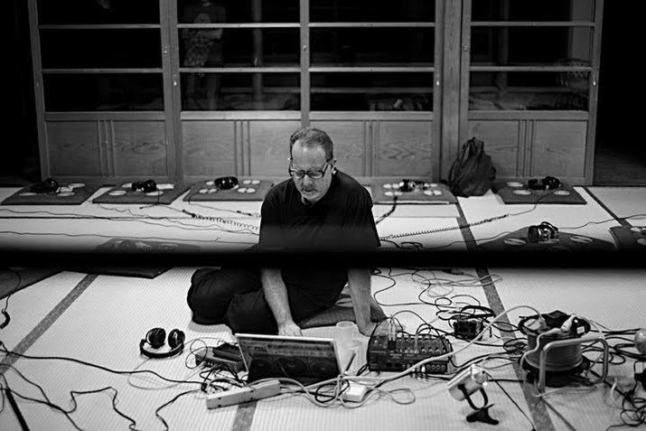 A black and white photograph of Carl Stone sitting on the floor surrounded by intertwining, curling wires. He looks down at a laptop in front of him. He wears all dark clothes and black glasses. Behind him, there are empty shelves. 