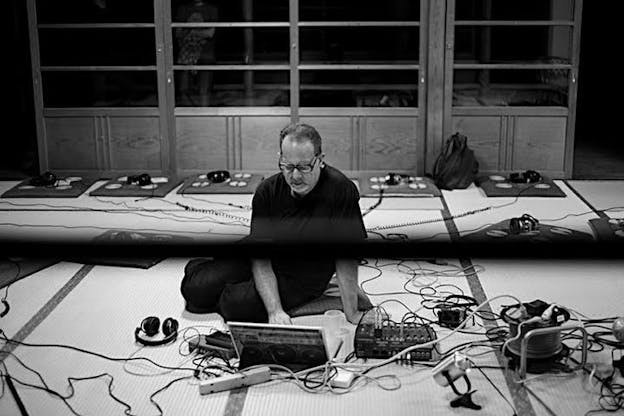 A black and white photograph of Carl Stone sitting on the floor surrounded by intertwining, curling wires. He looks down at a laptop in front of him. He wears all dark clothes and black glasses. Behind him, there are empty shelves. 
