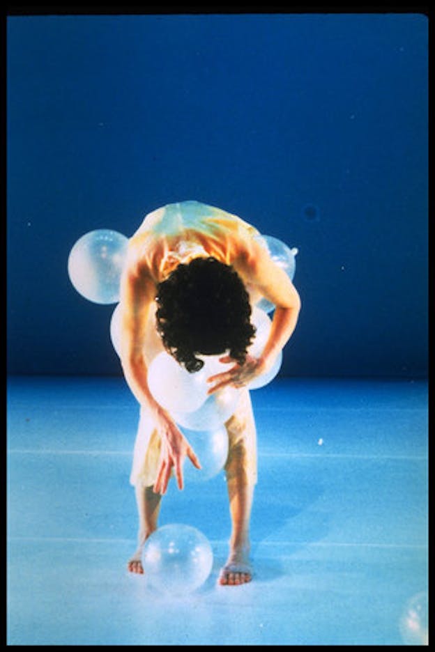 One person wearing light orange clothes bends forward in a bright blue space. They hold clear ballons by their torso which spill outwards and onto the floor. 
