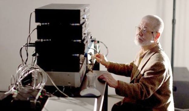 Yasunao Tone turns a knob on a sound machine in a bare space, He wears a tweed blazer and has a wire connected to his head. In this room, there are white walls, a metal desk, and five sound boxes stacked on top of it.
