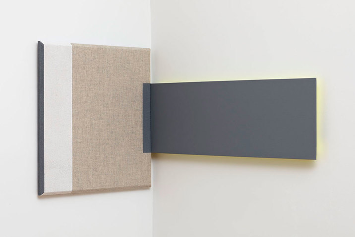 A square painting with a line of black on its left side, a thicker one in beige next to it and a bigger one in a brown-beige color stand on the left side of a wall. To its right on the wall a black rectangular shaped painting touches the other one. The square painting on the place where both meet has a black slot painted.