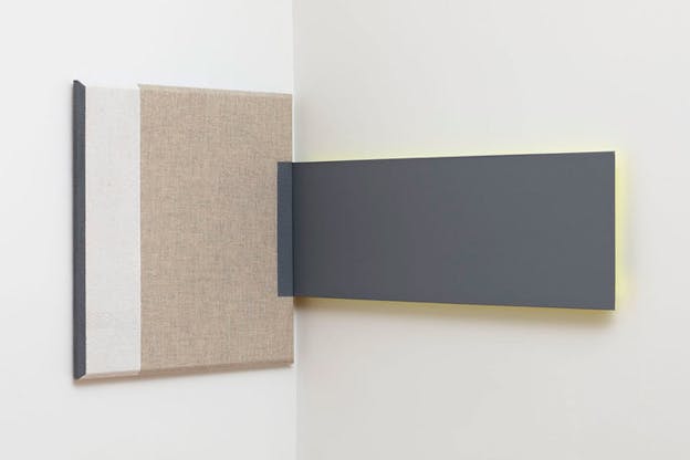 A square painting with a line of black on its left side, a thicker one in beige next to it and a bigger one in a brown-beige color stand on the left side of a wall. To its right on the wall a black rectangular shaped painting touches the other one. The square painting on the place where both meet has a black slot painted.