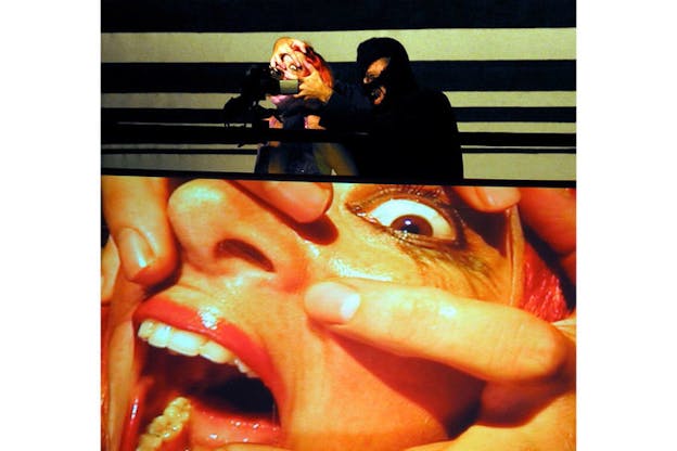 In the upper half of this image, a performer pulls at their face with their hands as another performer who is sheilded in shadows stands beside them. The performer pulling at their own face is projected onto a large screen on the lower half of this image.