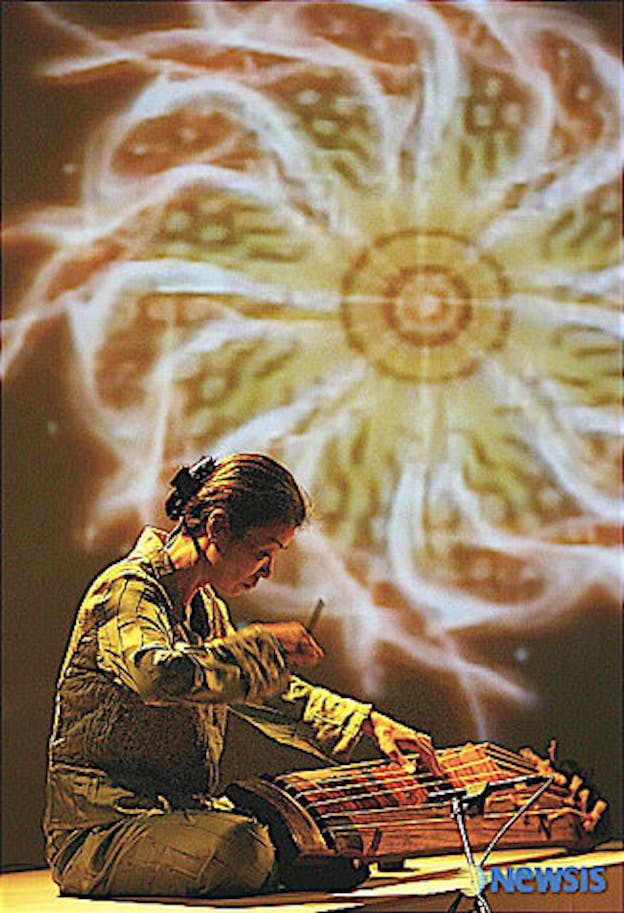 Jin Hi Kim sits in front of a komungo and plays it. She wears a green jumpsuit. A spiral of red and yellow is projected on the wall behind her.