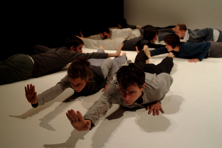 A performance still of several performers laying on their stomachs on a white floor, facing different directions. Each performer extends their right hand in front of them while keeping their left hand pressed to the floor besides their chests. 