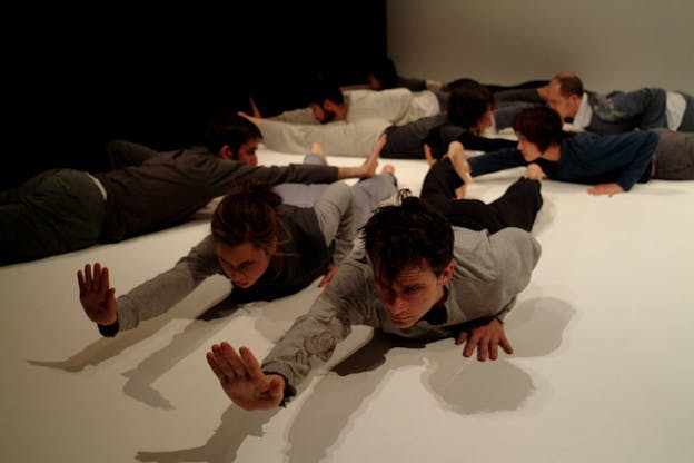 A performance still of several performers laying on their stomachs on a white floor, facing different directions. Each performer extends their right hand in front of them while keeping their left hand pressed to the floor besides their chests. 