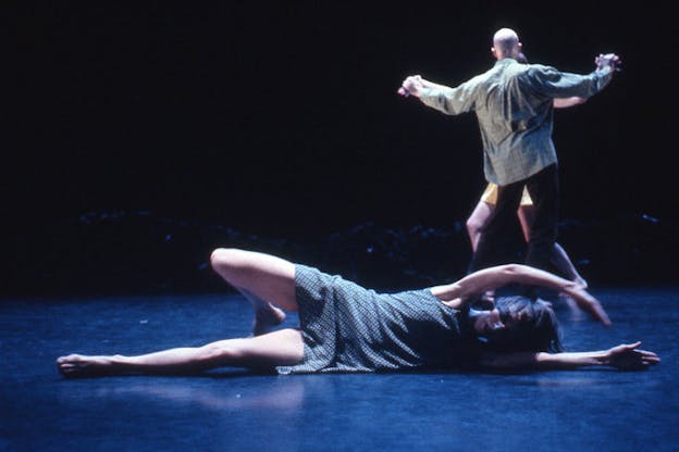 A performer wearing a short blue patterned dress lays on the floor of a dark stage with their arms above their head, their right leg bent at the knee and their left leg extended long on the ground. Two other performers dance together and hold hands in the background.