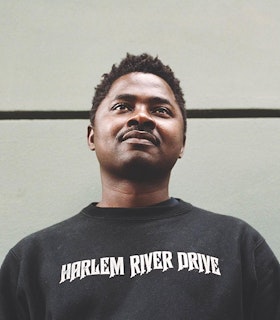 A low-angle portrait of Lamin Fofana, in front of a white background with one black horizontal line. The artist wears a black t-shirt that reads "Harlem River Drive."