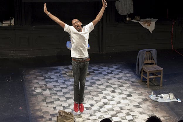 A person dressed in a white shirt, jeans and red shoes jumps on stage with their hands upwards in joy. The backheads of the audience can be seen staring towards the checkered stage floor.