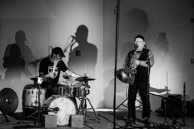 A black and white photograph of a person playing the drums and another playing the saxophone.