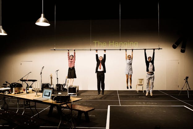 A performance still of four performers in street clothes hanging by their hands from a suspended horizontal rod. On the white wall behind them, there is a projection that reads 
