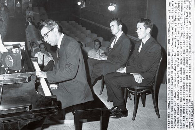 Black and white photograph of a pianist playing in a theater. Two people are seated in chairs directly behind the performer, a small audience is scattered in various seats. 