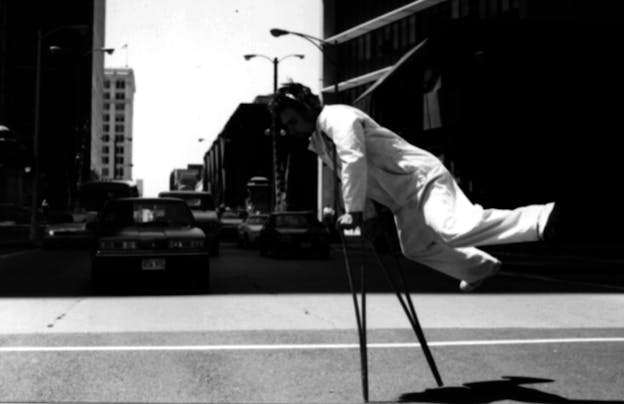 Bill Shannon rests on his arms on crutches in front of him as the lower half of his body is held upwards behind him nearly horizontal to the ground. He stands in a city street. 