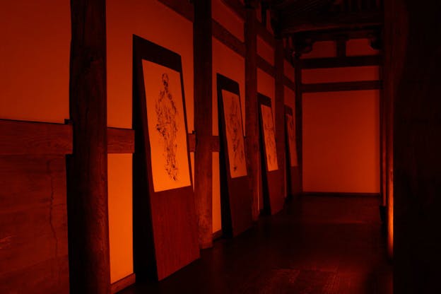 Within a dark, red hued room, four wooden panels lean against the wall. On each wooden panel is a white sheet of paper with ink line drawings on it. The wall itself is primarily white and segmented with wooden rods and columns which make a grid.