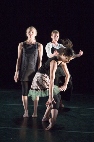 Three performers wearing mismatched outfits are posed differently in front of a black backdrop. The first performer extends and arches their right foot while reaching downward with both of their hands. The second performer stands upright behind the first performer while staring upward. In the very back, the third performer bends their knees and leans back while holding their fists in the front. 