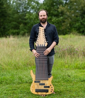 Zach stands on green grass with wildflowers and trees in the background. He is wearing a dark button-down shirt and holds a 17-string electric bass upright with his hands on its neck and its body resting on the ground.