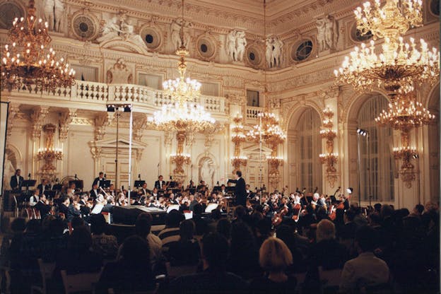 An orchestra plays while an audience watches seated down. Shining gold chandeliers hang from the white ceiling which is decorated with intricate flower carvings and small Roman inspired statues.