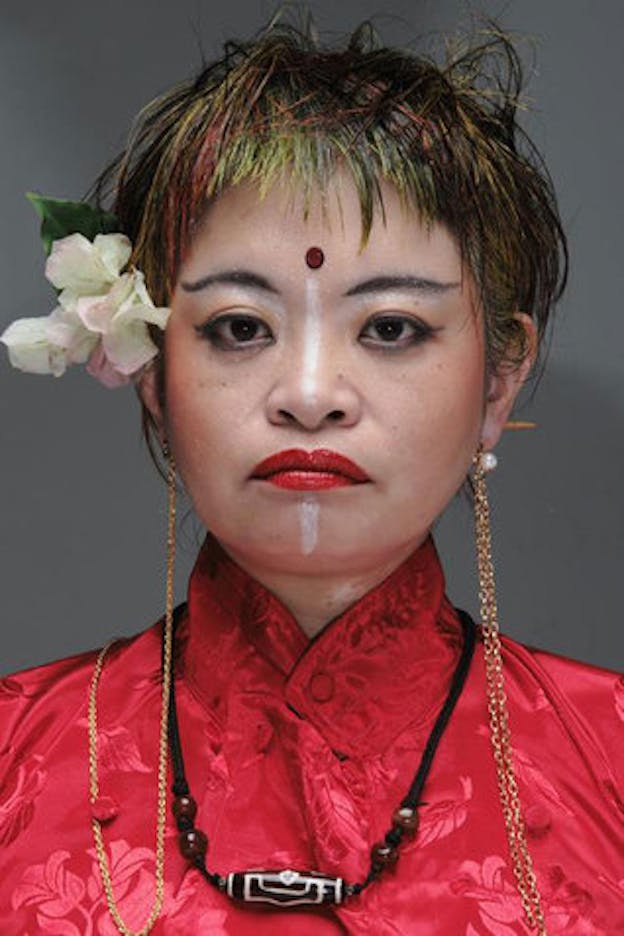 Nexus6 performs in a red top with white flowers tucked behind her right ear. She wears eyeliner and red lipstick. 