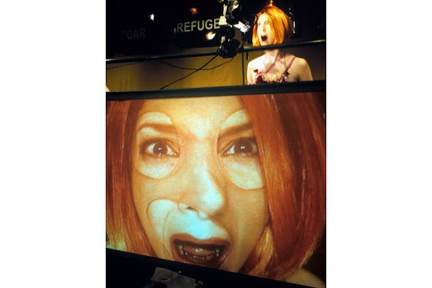 A performer wearing a sheer mask with holes cut out for their eyes and mouth looks into a camera appearing distressed. A large screen on the bottom half of this image projects that performer.