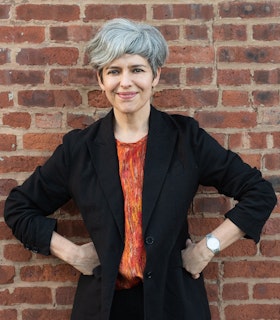 A portrait of Monica de la Torre, smiling and standing in front of a brick wall with her hands at her hips. She has short grey hair and wears a black blazer, an orange and red abstract patterned shirt, and a silver watch.