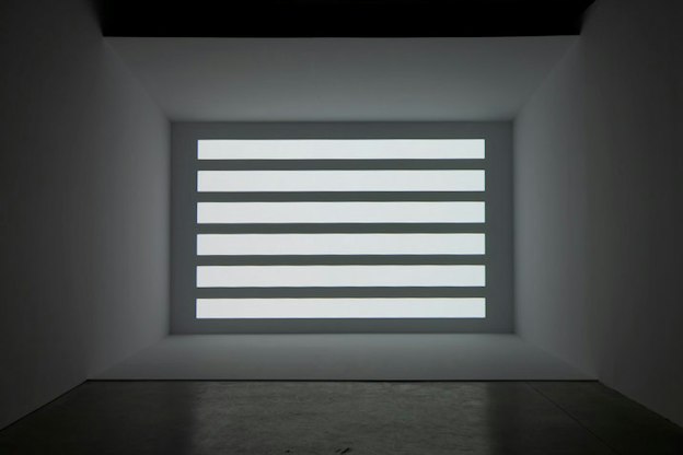 At the far wall of a dark grey room, there is a grey wall with various horizontal bars of light projected onto it.