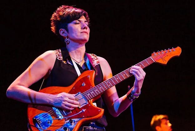 Ava Mendoza plays a wood grain patterned electric guitar on a stage, lit by orange and blue lights. Visible from the hips up, their body is turned slightly to the right, and their eyes are closed. They are wearing a black sleeveless button up top with leopard print highlights on the shoulders, a watch and leather band bracelet on their left wrist, a chain necklace, and a pair of round, dangling earrings.
