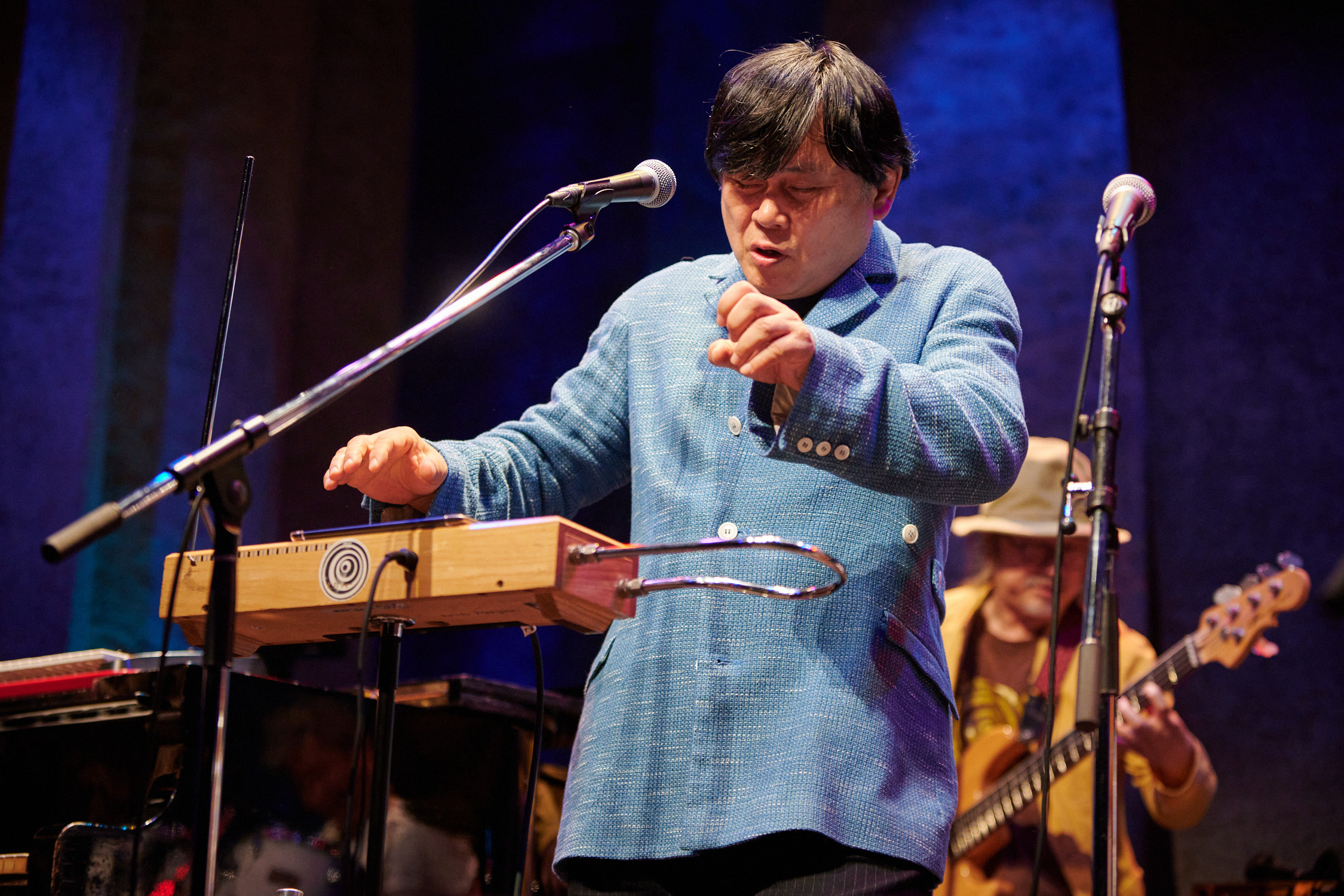 Koichi Makigami is centered on a stage with a blue backdrop. A bass player is pictured behind him and slightly out of focus. He is turned slightly to the left with his eyes closed and plays a theremin while vocalizing into a microphone. He is wearing a woven, light blue blazer with white buttons. 