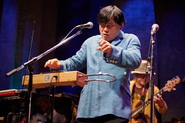Koichi Makigami is centered on a stage with a blue backdrop. A bass player is pictured behind him and slightly out of focus. He is turned slightly to the left with his eyes closed and plays a theremin while vocalizing into a microphone. He is wearing a woven, light blue blazer with white buttons. 