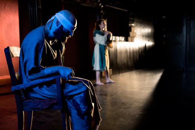 In a narrow dimly-lit room, a person dressed in rags with their eyes blindfolded, their body hued with blue light sits on a chair with their back hunched. In the distance, a barefoot person in a dress stands blurred.