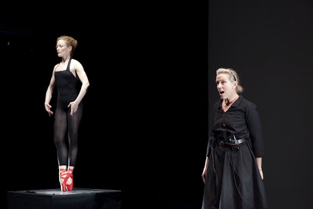 A figure clad in black stands on a stage speaking, next to her another perfromer dressed in black with red pointe shoes stands in a releve position with her weight supported by her toes.