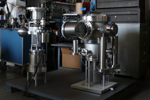 A silver machine rests on a black wooden board in front of shelving units and a cluttered blue table. The machine is composed of numerous cylinders. 