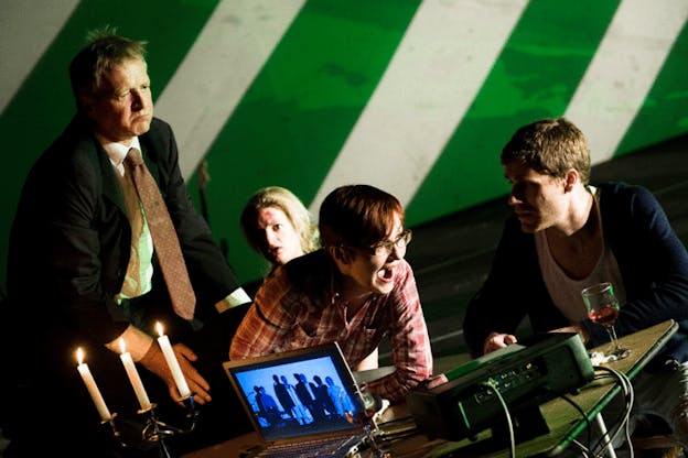 A performance still of four performers clustered around a table filled with a lit candelabra, sound equipment, glasses of red wine, and a laptop which shows an image of people standing and looking intently ahead. One performer leans forward on the table with an exuberant expression on their face while the three performers around them look concerned. The wall in the background is painted with diagonal white and green stripes. 