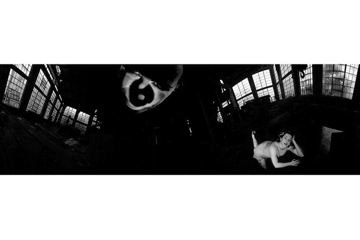 A black and white panoramic image has in the center of it closest to the viewer a face half covered by shadows. Behind it another figure lies on the floor their angled hand supporting their head. 