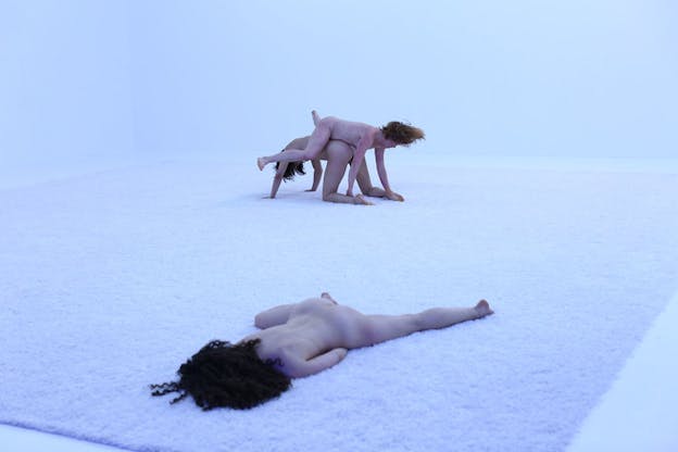 Two naked figures stand on top of each other in all fours as a third one closest to the viewer lies down with their front on the white carpet, their face covered by their brown hair.