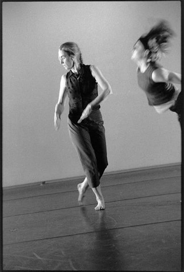 Two performers dance in a dark space. One wears a dark outfit and a braid and stands in the center, resting on one leg and looking downwards. The other performer is visible only from the waist up. They bend forward dramatically, throwing their long hair upwards in the process.